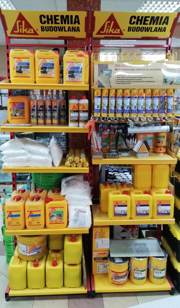 Ekspozycja chemii budowlanej marki Sika na żółtym regale sklepowym, widoczne kanistry, butelki, kartusze z uszczelniaczami i klejami, oraz worki z materiałami sypkimi.