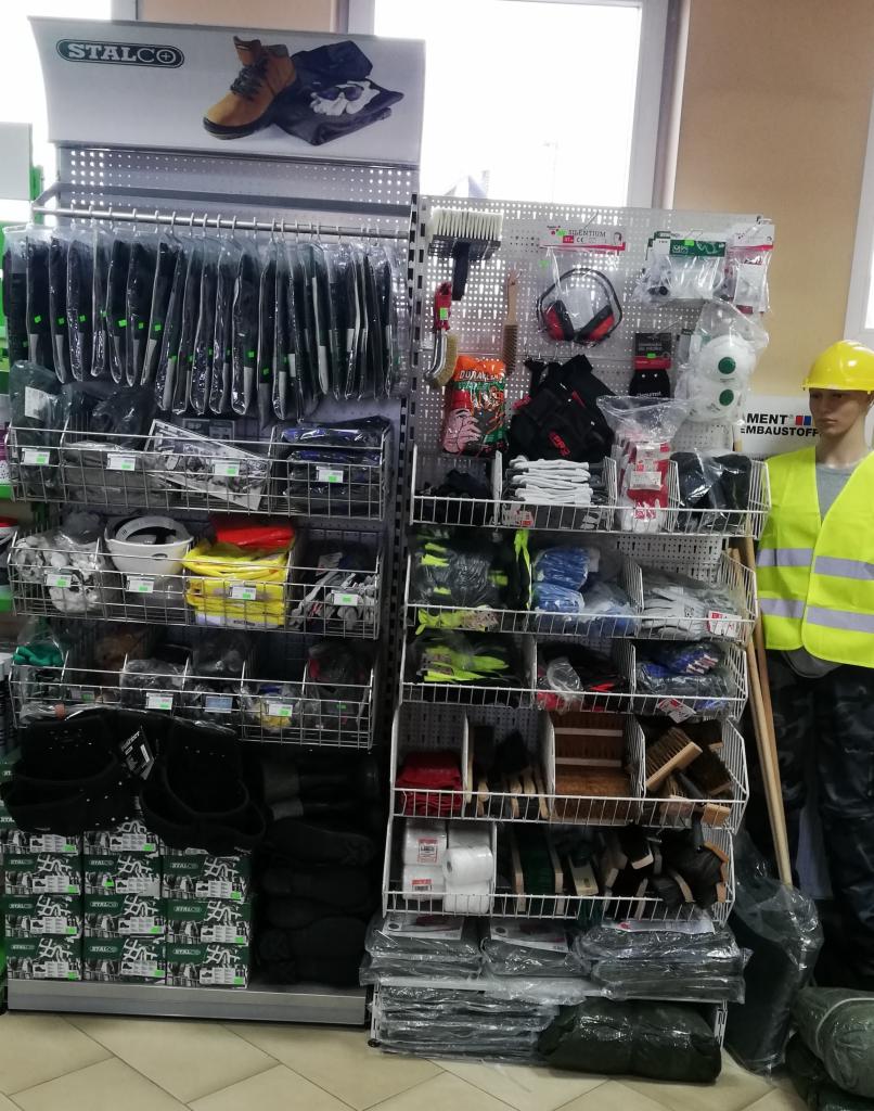 Ekspozycja w sklepie z artykułami BHP i narzędziami: odzież robocza, obuwie, rękawice, maski, słuchawki, pędzle i inne akcesoria na metalowych regałach. Manekin w żółtej kamizelce odblaskowej...