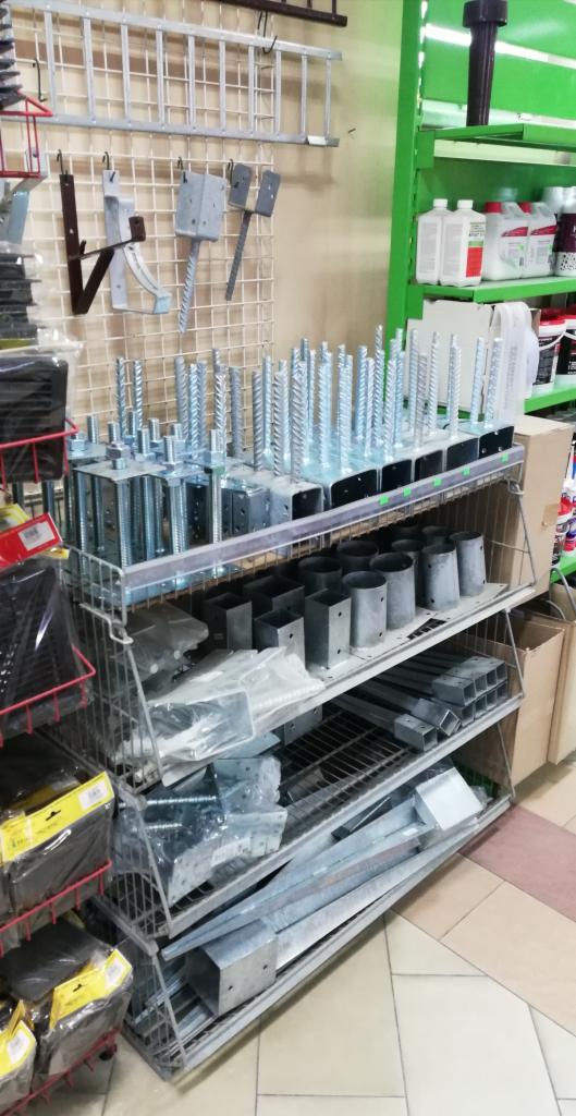 Metalowe elementy konstrukcyjne, w tym śruby kotwiące, uchwyty i profile, ułożone na półkach w sklepie budowlanym.