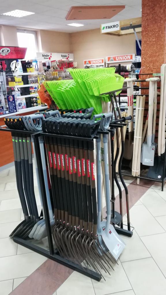 Ekspozycja w sklepie budowlanym: rzędy grabi, szpadli i wideł z plastikowymi uchwytami, ustawione na metalowym stojaku, z zielonymi koszami budowlanymi w tle.