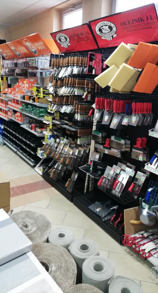 Półki w sklepie budowlanym z narzędziami do prac wykończeniowych, w tym szpachle z drewnianymi i czerwonymi rączkami, gąbki do czyszczenia oraz wałki z włókna na podłodze.