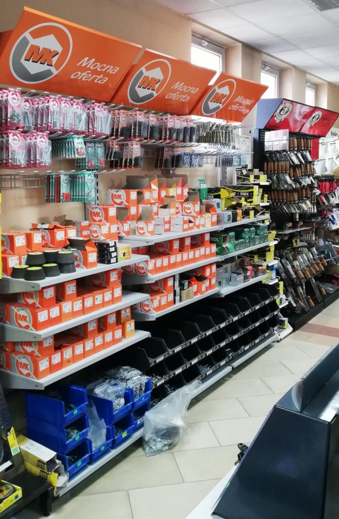 Półki w sklepie budowlanym z materiałami montażowymi, narzędziami i akcesoriami, widoczne pomarańczowe pudełka i narzędzia na wieszakach.