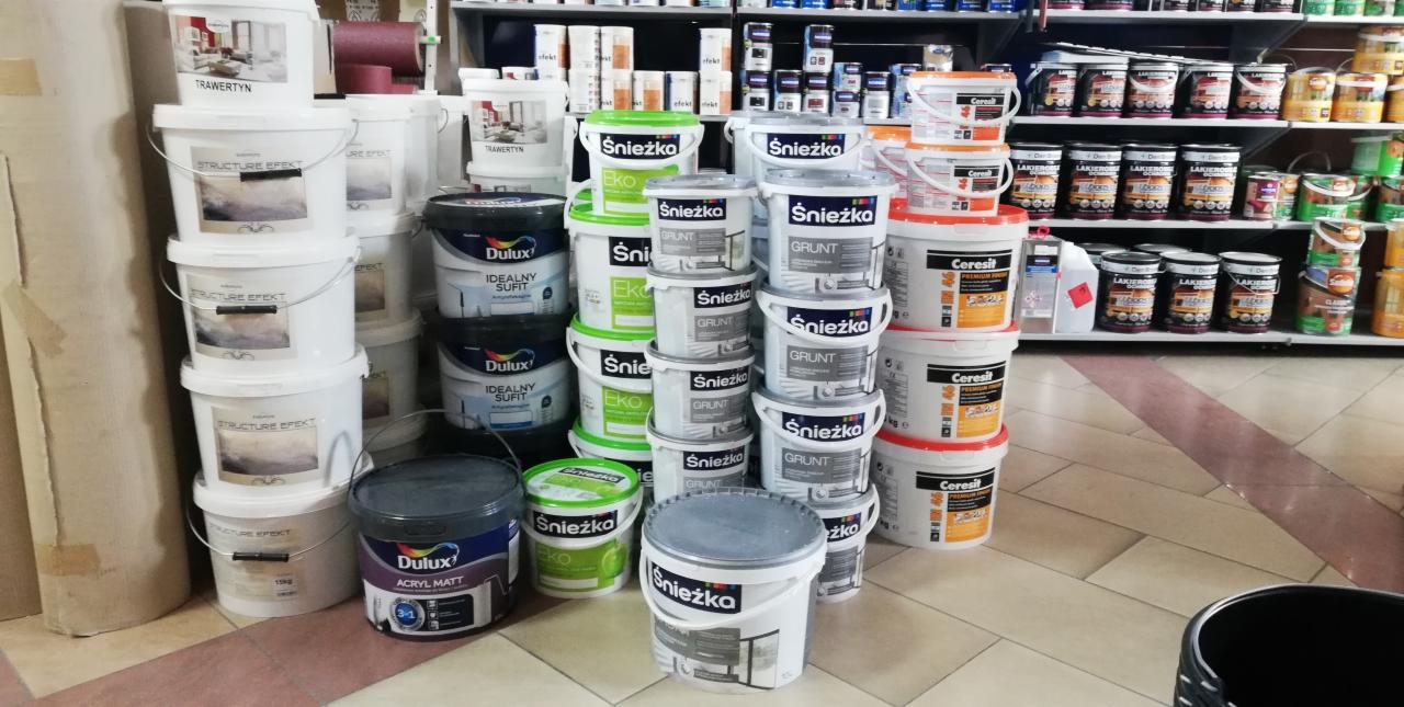 Różnorodne farby i grunty różnych marek (Śnieżka, Dulux, Ceresit) ustawione w rzędach na podłodze sklepu budowlanego, widoczne także półki z innymi produktami.
