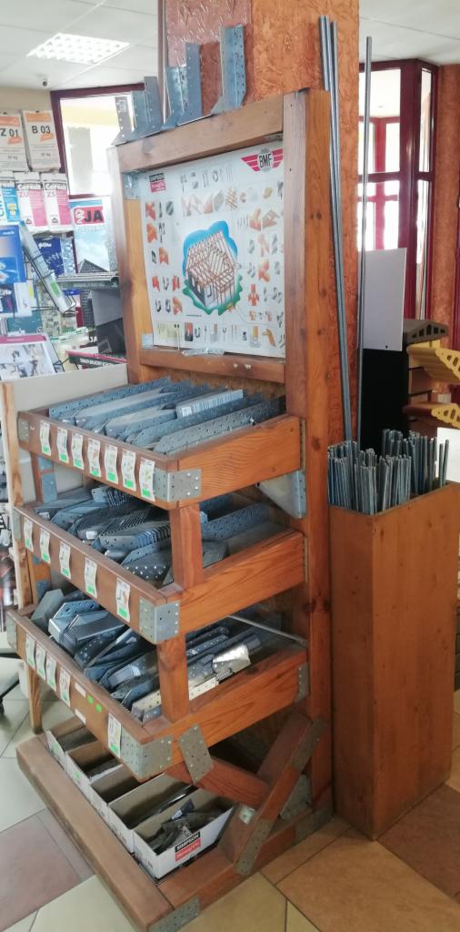 Ekspozycja w sklepie z drewnianym regałem prezentującym metalowe złącza i akcesoria do konstrukcji dachowych, obok pionowy pojemnik z metalowymi prętami.