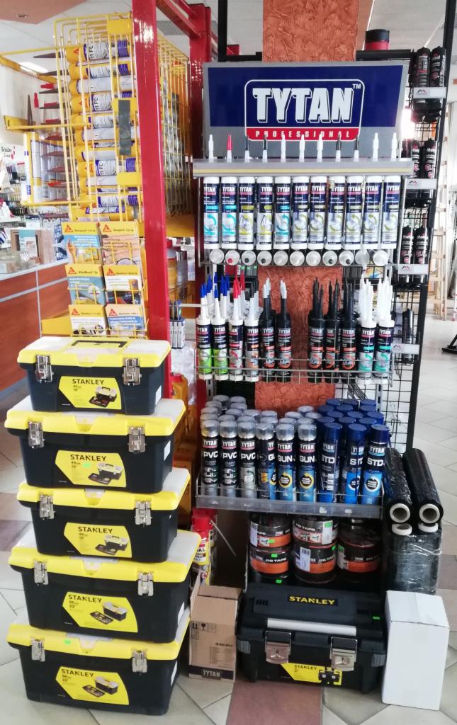 Ekspozycja w sklepie budowlanym: pełna półka z produktami Tytan Professional, obok stos skrzynek narzędziowych Stanley, widok z perspektywy klienta.