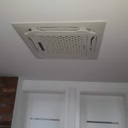 Kasetonowa jednostka klimatyzacyjna zamontowana w suficie podwieszanym, widoczna z dołu, z fragmentem białych drzwi i ściany z cegły w tle.