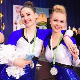 Dwie uśmiechnięte cheerleaderki w błyszczących niebiesko-białych strojach pozują na tle sceny, trzymając pompony i smartfon w kształcie rekina. Na szyjach mają medale za Mistrzostwa Polski...