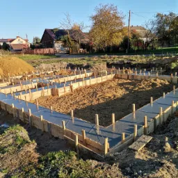 Wylany fundament pod dom jednorodzinny z drewnianym szalunkiem, widok z góry, w tle zabudowa mieszkalna i błękitne niebo.