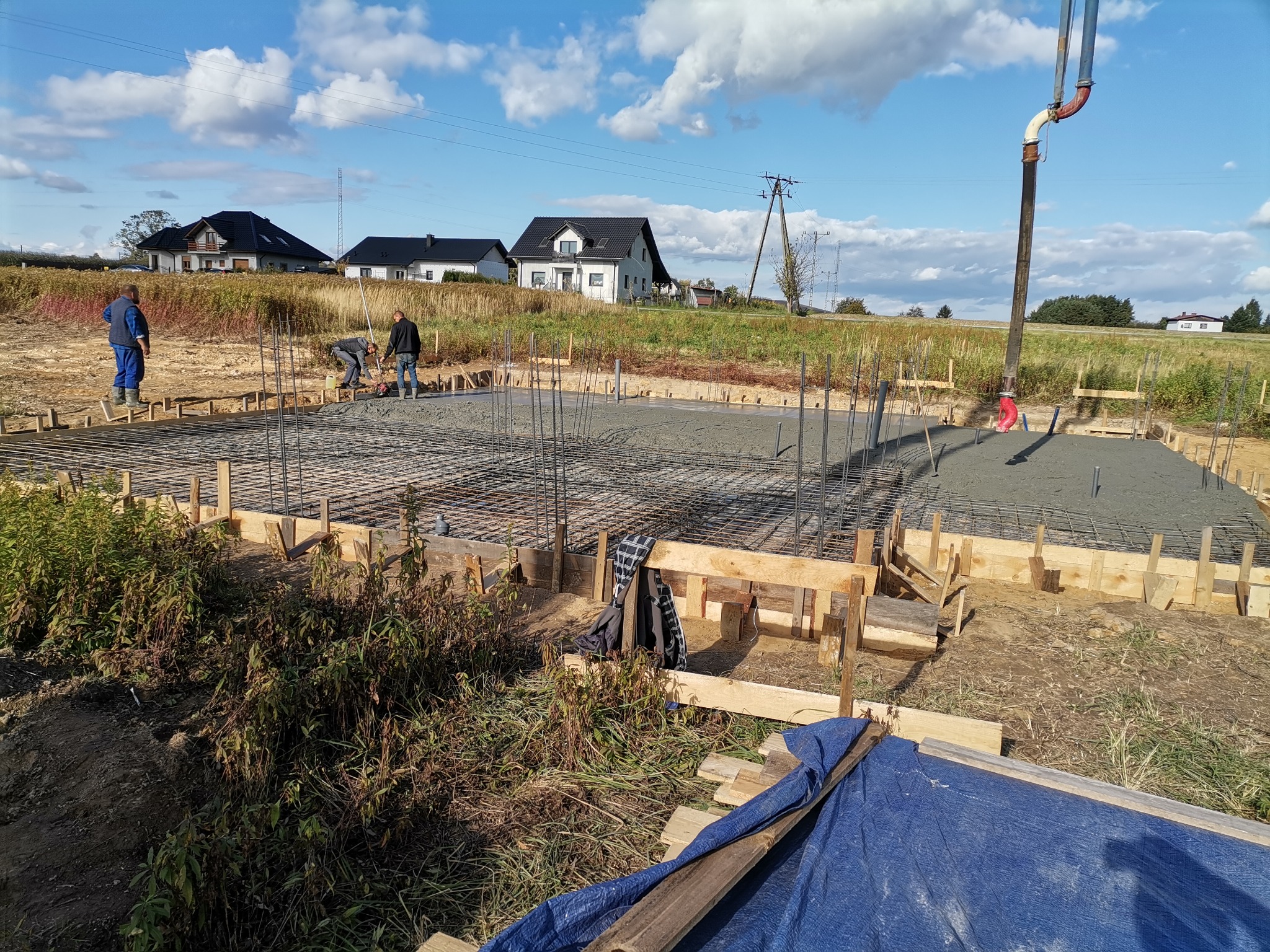 Wylewanie fundamentów pod dom jednorodzinny z użyciem pompy do betonu; widoczne stalowe zbrojenie i drewniane szalunki, w tle domy w budowie.