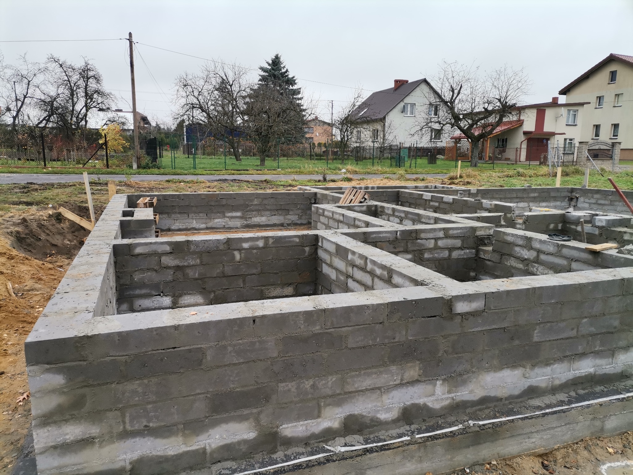 Wykopane fundamenty z betonu komórkowego pod budynek mieszkalny jednorodzinny, widok na działkę z zabudową mieszkalną w tle, pochmurny dzień.