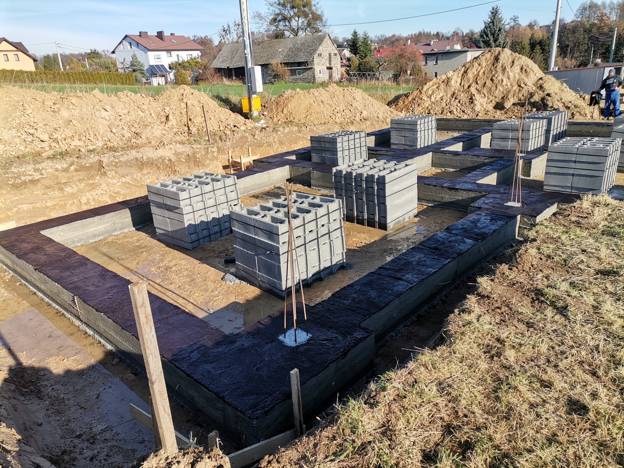 Fundamenty domu w trakcie budowy, z izolacją przeciwwilgociową i ułożonymi bloczkami betonowymi, widoczne wykopane rowy i usypana ziemia w tle.