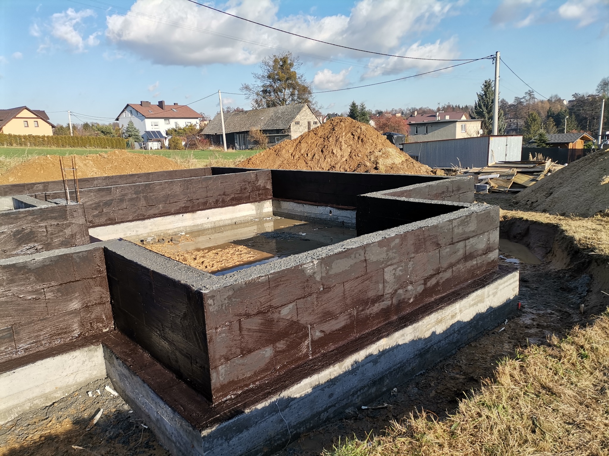 Fundamenty domu z bloczków betonowych, zabezpieczone brązową izolacją bitumiczną, zalane wodą, z widoczną stertą ziemi w tle i zabudową jednorodzinną.