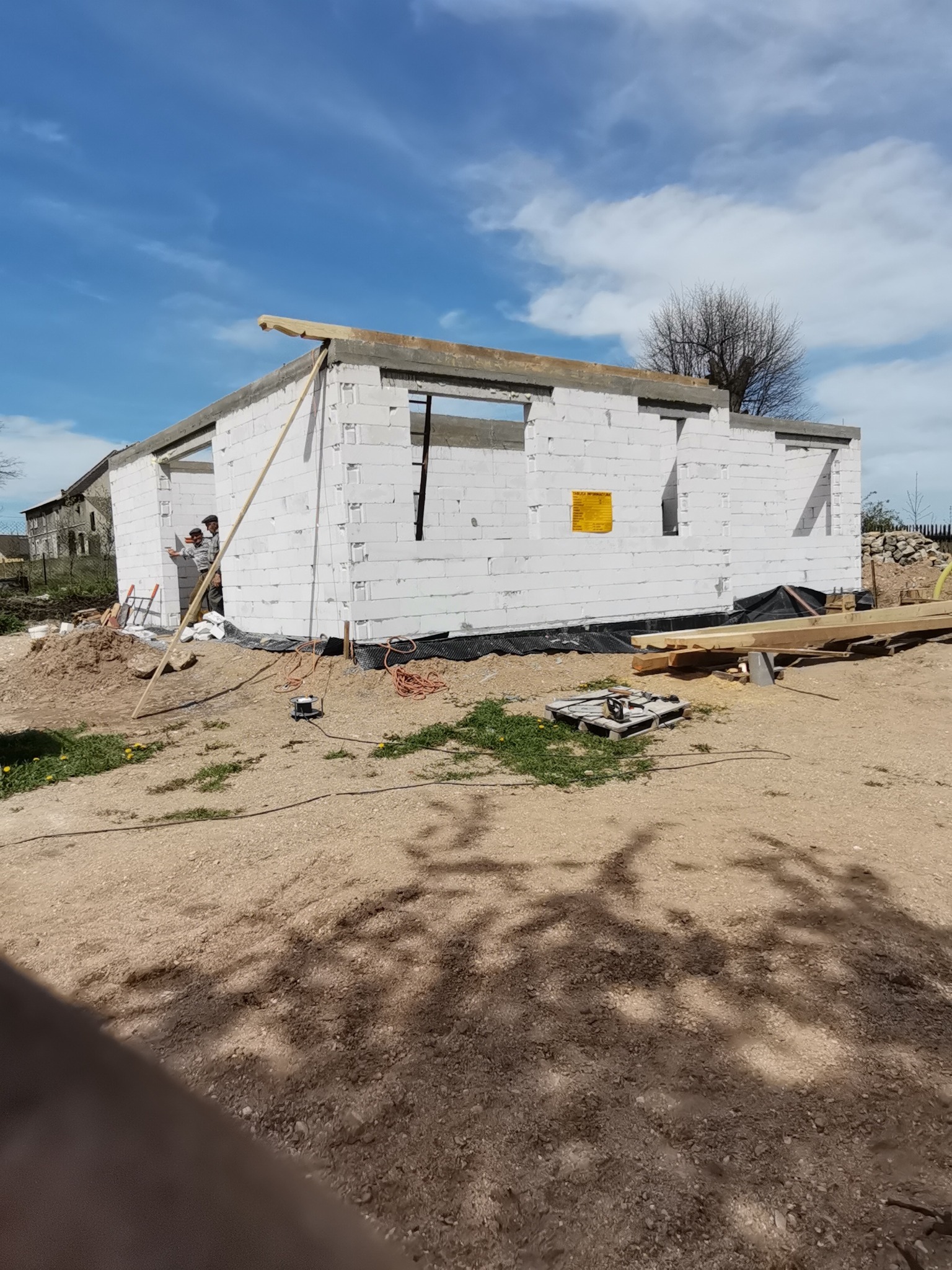 Wznoszenie ścian parterowego budynku z białych bloczków, widoczni dwaj pracownicy przy pracy, przygotowane materiały budowlane na placu budowy.