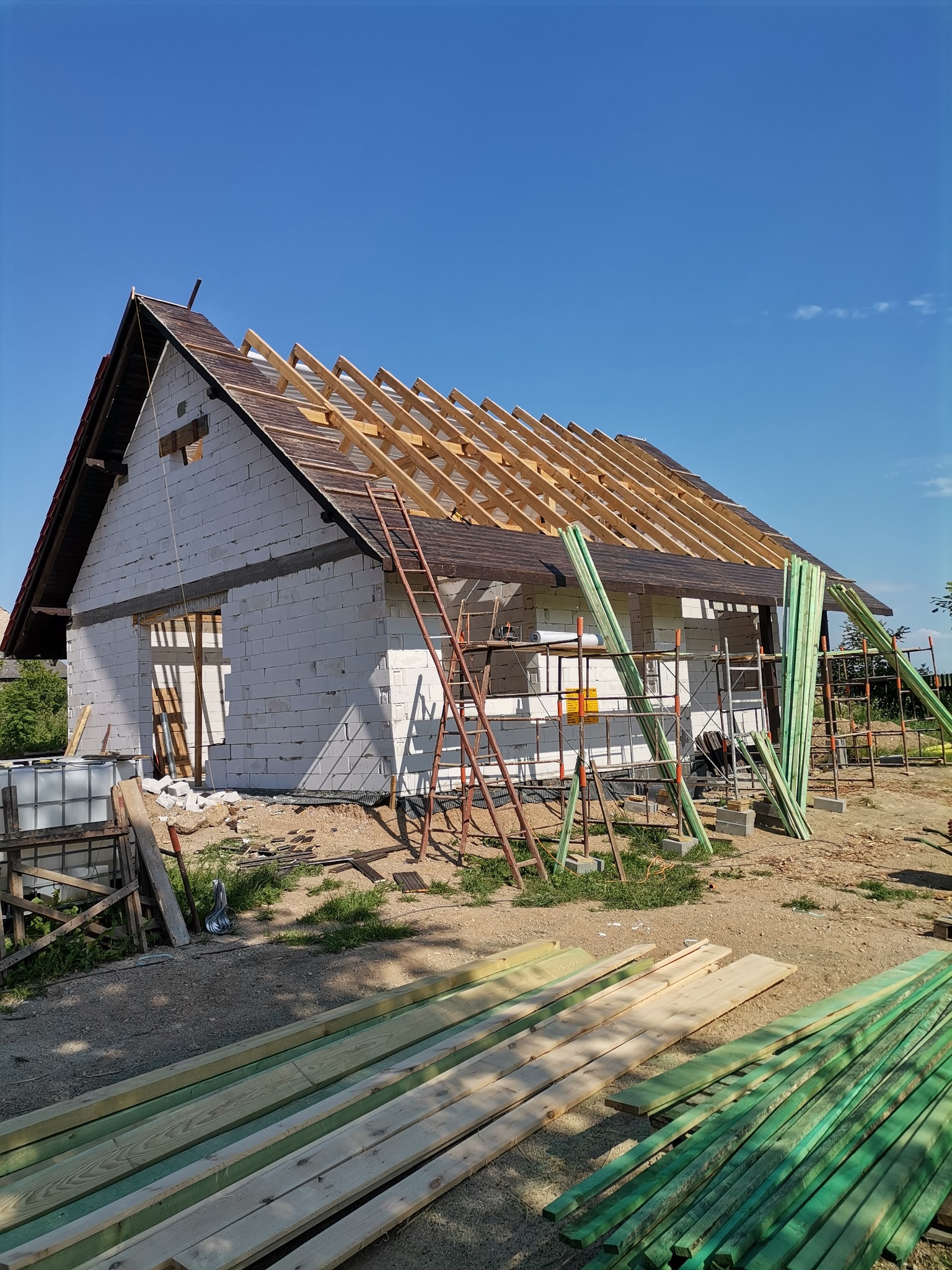 Murowany dom w trakcie budowy, widoczne ściany z białych bloczków, częściowo pokryty dach z drewnianą konstrukcją, rusztowania i stosy impregnowanych desek na pierwszym planie.