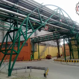 Konstrukcja stalowa nad drogą z rurociągami, znak ograniczenia wysokości 5.8m. W tle budynki przemysłowe i mała ciężarówka. Ujęcie z dołu na zewnątrz.