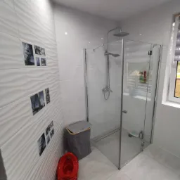 Nowoczesna łazienka z kabiną prysznicową typu walk-in, białe płytki z falistym wzorem, dekoracyjne zdjęcia na ścianie, czerwony nocnik dziecięcy i kosz na pranie.