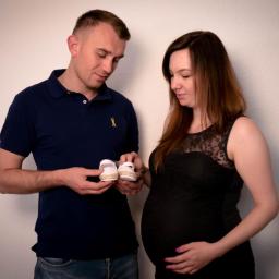 Crazy Team Media - Przyszli rodzice spoglądają na małe białe buciki dla dziecka, kobieta w czarnej sukience trzyma dłonie na brzuchu.