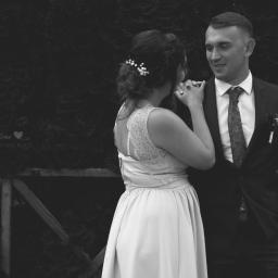 Crazy Team Media - Czarno-białe zdjęcie pary młodej: panna młoda w sukni z koronkowymi plecami i upiętymi włosami z ozdobami, trzymająca dłoń pana młodego w garniturze z butonierką, na tle ciemnej zieleni.