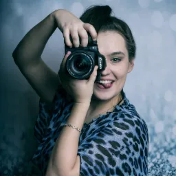 Uśmiechnięta fotografka z wyciągniętym językiem celuje aparatem Canon z obiektywem zoom, pozując na tle abstrakcyjnego, błękitnego tła.