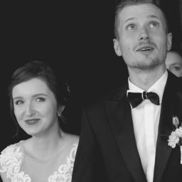 Crazy Team Media - Czarno-białe zdjęcie młodej pary: panna młoda uśmiecha się w lewo, pan młody patrzy w górę, oboje stoją na tle ciemnego wejścia.