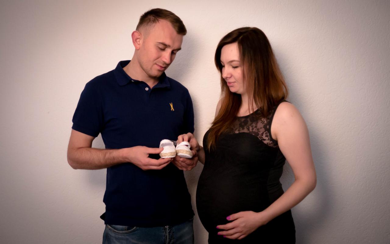 Przyszli rodzice spoglądają na małe białe buciki dla dziecka, kobieta w czarnej sukience trzyma dłonie na brzuchu.