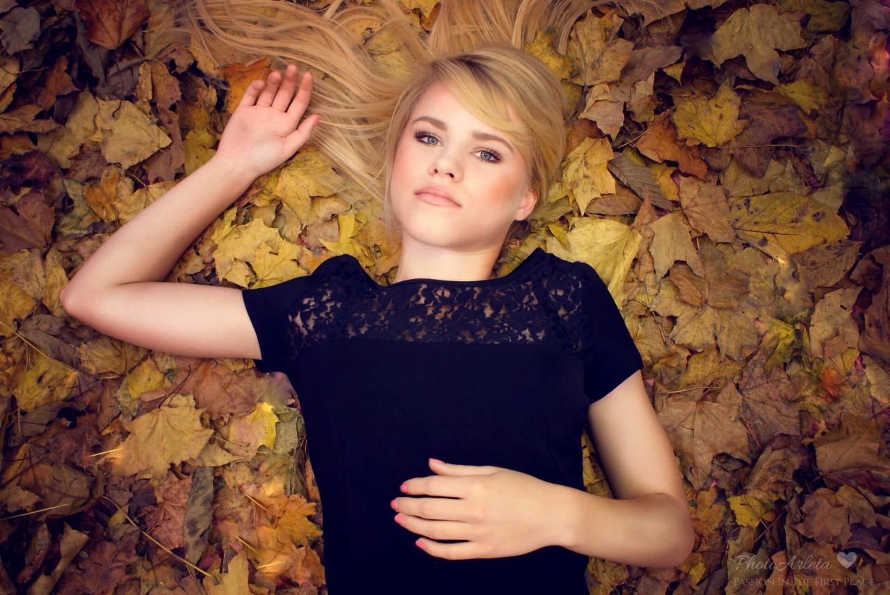 Młoda kobieta w czarnej sukience leży na dywanie z opadłych, jesiennych liści, z uniesioną ręką i delikatnym uśmiechem, patrząc w górę.