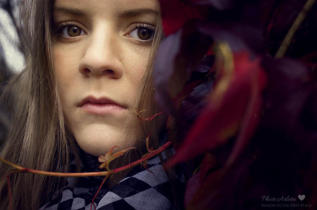 Portret młodej kobiety z brązowymi oczami, otoczonej jesiennymi, czerwonymi liśćmi i gałązkami, ubrana w szaro-czarną szachownicę.