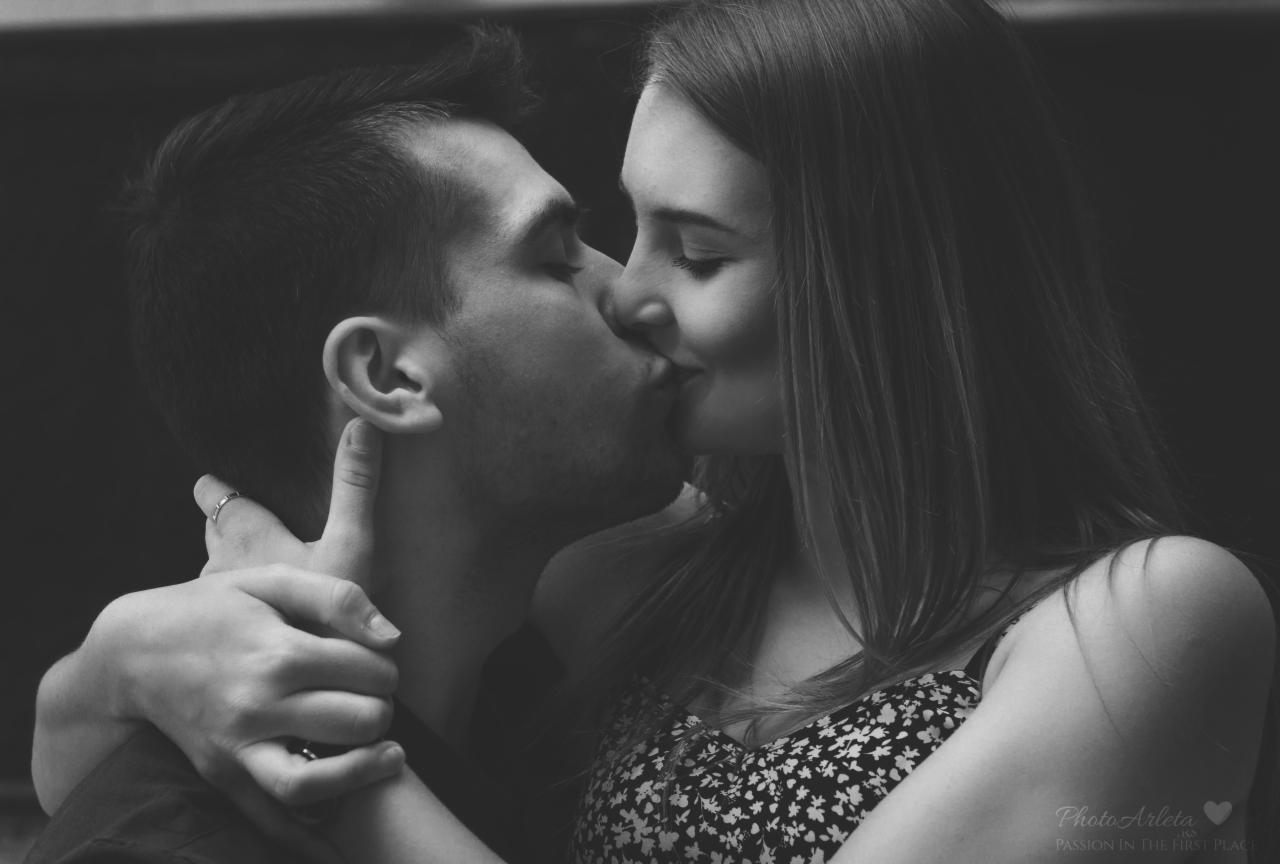 Czarno-białe zbliżenie całującej się pary; dłoń mężczyzny z pierścionkiem obejmuje szyję kobiety w kwiecistej sukience.