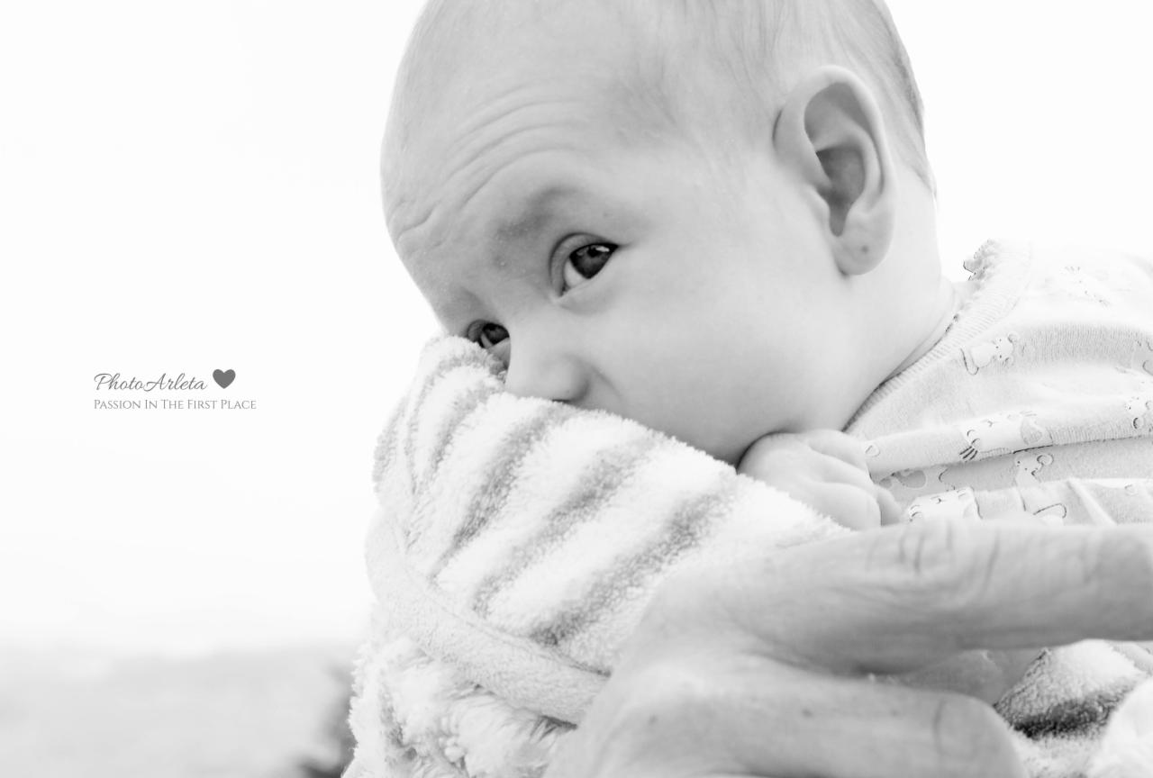 Czarno-białe zbliżenie noworodka trzymającego w dłoniach pasiasty kocyk, zamyślony wyraz twarzy, widoczne zmarszczki na czole, w tle delikatny krajobraz i logo fotografa.