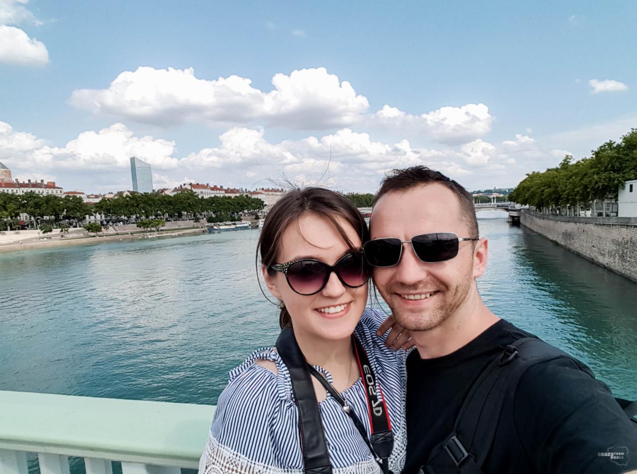 Uśmiechnięta para w okularach przeciwsłonecznych pozuje na moście z widokiem na rzekę i panoramę miasta z wieżowcem w tle.