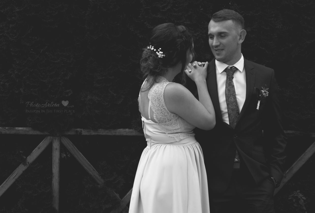 Czarno-białe zdjęcie pary młodej: panna młoda w sukni z koronkowymi plecami i upiętymi włosami z ozdobami, trzymająca dłoń pana młodego w garniturze z butonierką, na tle ciemnej zieleni.