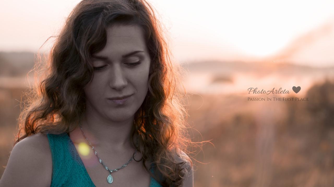 Portret kobiety z kręconymi włosami, spuszczonym wzrokiem i srebrnym naszyjnikiem na tle rozmytego krajobrazu o zachodzie słońca.