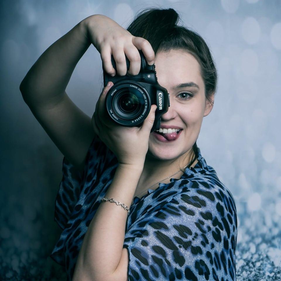 Uśmiechnięta fotografka z wyciągniętym językiem celuje aparatem Canon z obiektywem zoom, pozując na tle abstrakcyjnego, błękitnego tła.