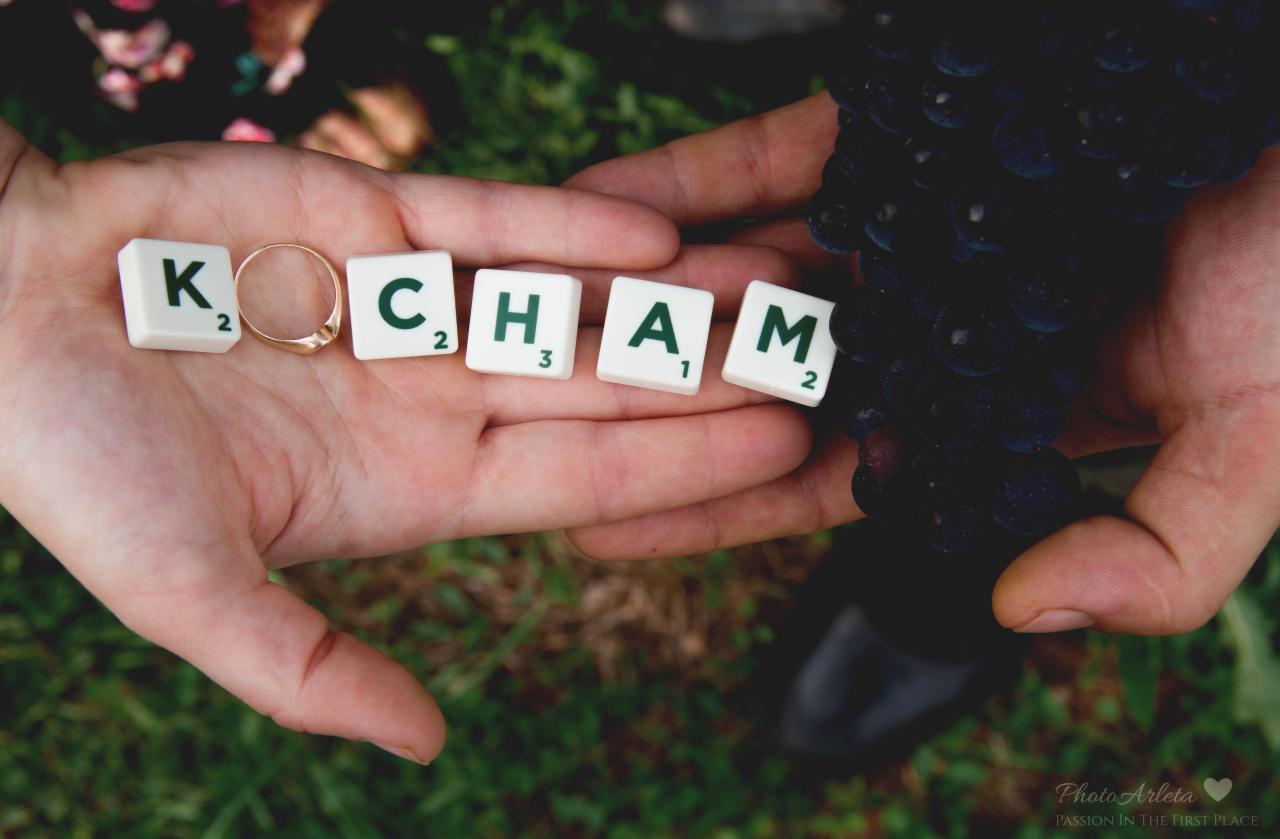 Dłonie trzymające litery z gry Scrabble układające się w słowo 'KOCHAM', z obrączką zamiast litery 'O' i kiścią ciemnych winogron w tle na zielonej trawie.