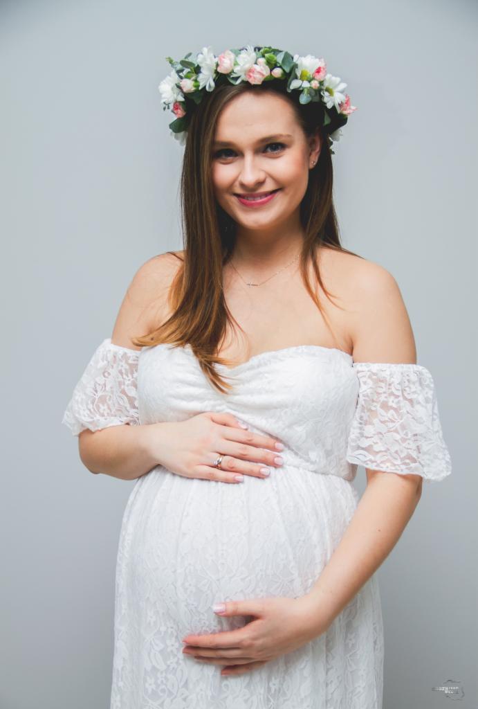Uśmiechnięta kobieta w ciąży w białej koronkowej sukience z odkrytymi ramionami i wiankiem z białych i różowych kwiatów na głowie, trzyma ręce na brzuchu.