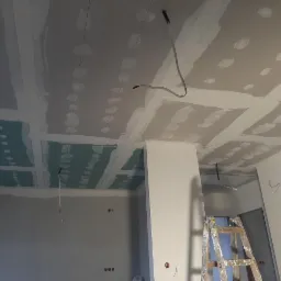 Montaż sufitu podwieszanego z płyt kartonowo-gipsowych, widoczne łączenia szpachlowane, wystające przewody elektryczne, drabina malarska w pomieszczeniu.
