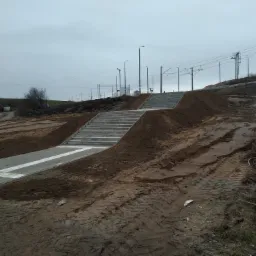 Betonowe schody i chodnik z białymi pasami dla niewidomych prowadzące na wzgórze, otoczone świeżo rozkopaną ziemią i śladami opon, w tle słupy wysokiego napięcia.