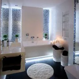 Nowoczesna łazienka z podświetlaną wanną, umywalką nablatową i lustrem w mozaikowej ramie, dekoracyjna mozaika na ścianach, okrągły puszysty dywanik na ciemnej podłodze.