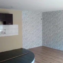 Wykończone wnętrze pokoju z panelami podłogowymi, ścianą pokrytą dekoracyjnym tynkiem imitującym kamień oraz częściowo widoczną zabudową kuchenną z białymi płytkami z motywem kwiatowym.