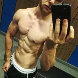 Mężczyzna z wyraźnie zarysowanymi mięśniami brzucha i ramion robi selfie w szatni z niebieskimi szafkami.