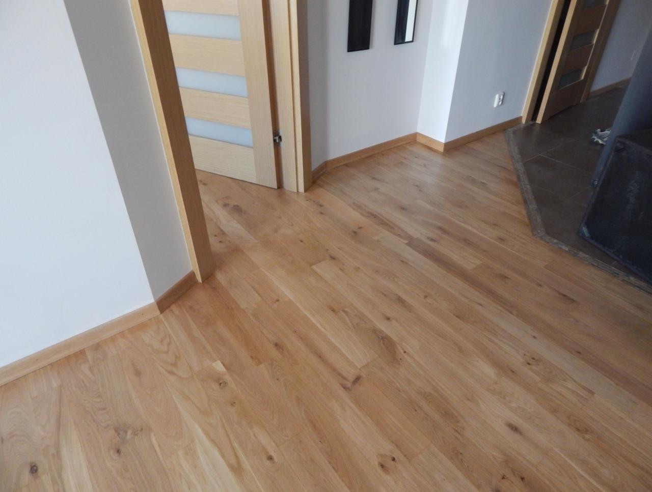 Wycyklinowana podłoga parkietowa z jasnego drewna w korytarzu, widoczne drzwi z przeszkleniami i białe ściany z listwami przypodłogowymi.