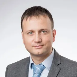 Portret mężczyzny w szarej marynarce i błękitnym krawacie na jasnoszarym tle, z delikatnym uśmiechem.