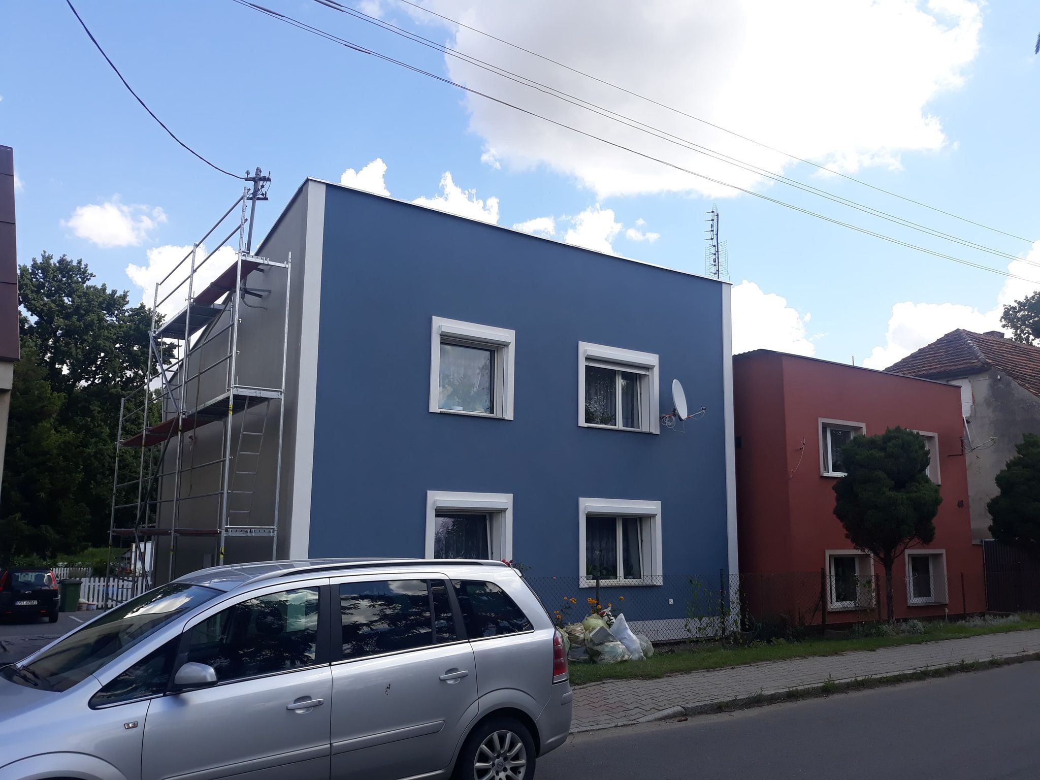Dwukondygnacyjny dom w trakcie ocieplania, widoczne rusztowania, elewacja w kolorze niebieskim, obok dom z elewacją w kolorze ceglastym, na pierwszym planie srebrny samochód.