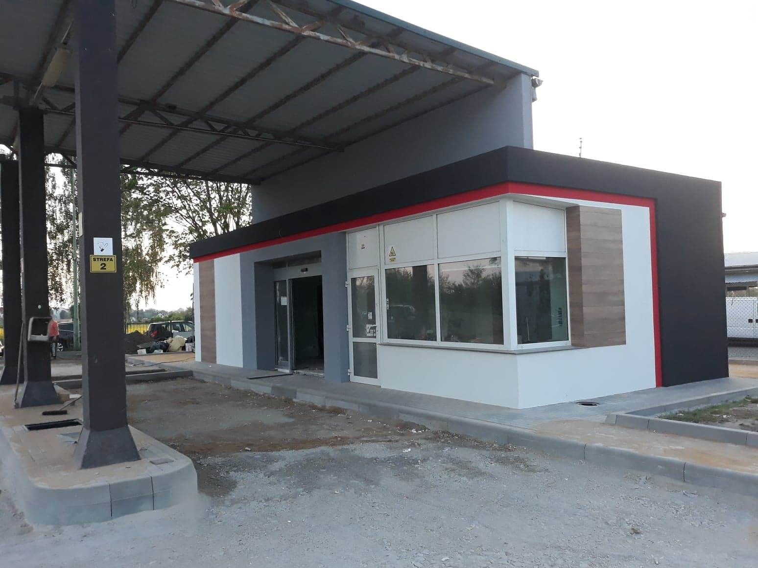 Nowoczesny budynek z oknami i automatycznymi drzwiami wejściowymi, wykończony w kolorach szarości, bieli i czerni z czerwoną linią akcentującą, w trakcie prac wykończeniowych na zewnątrz.