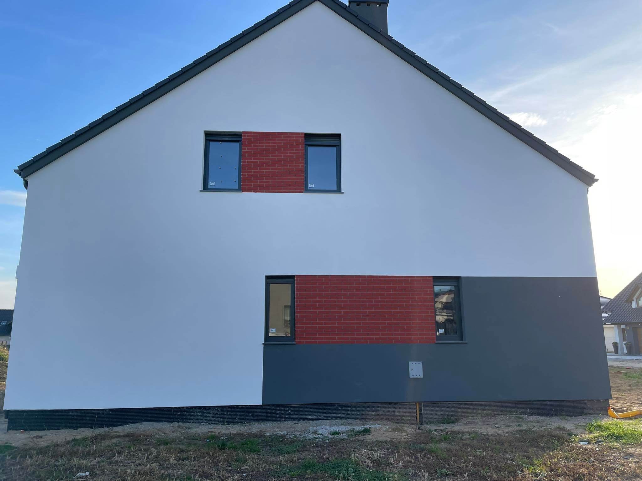 Elewacja domu z fragmentami wykończonymi czerwoną cegłą klinkierową pomiędzy oknami, biała górna część ścian i szara dolna, widoczne okna z ciemnymi ramami, podbitka dachu w kolorze grafitowym.