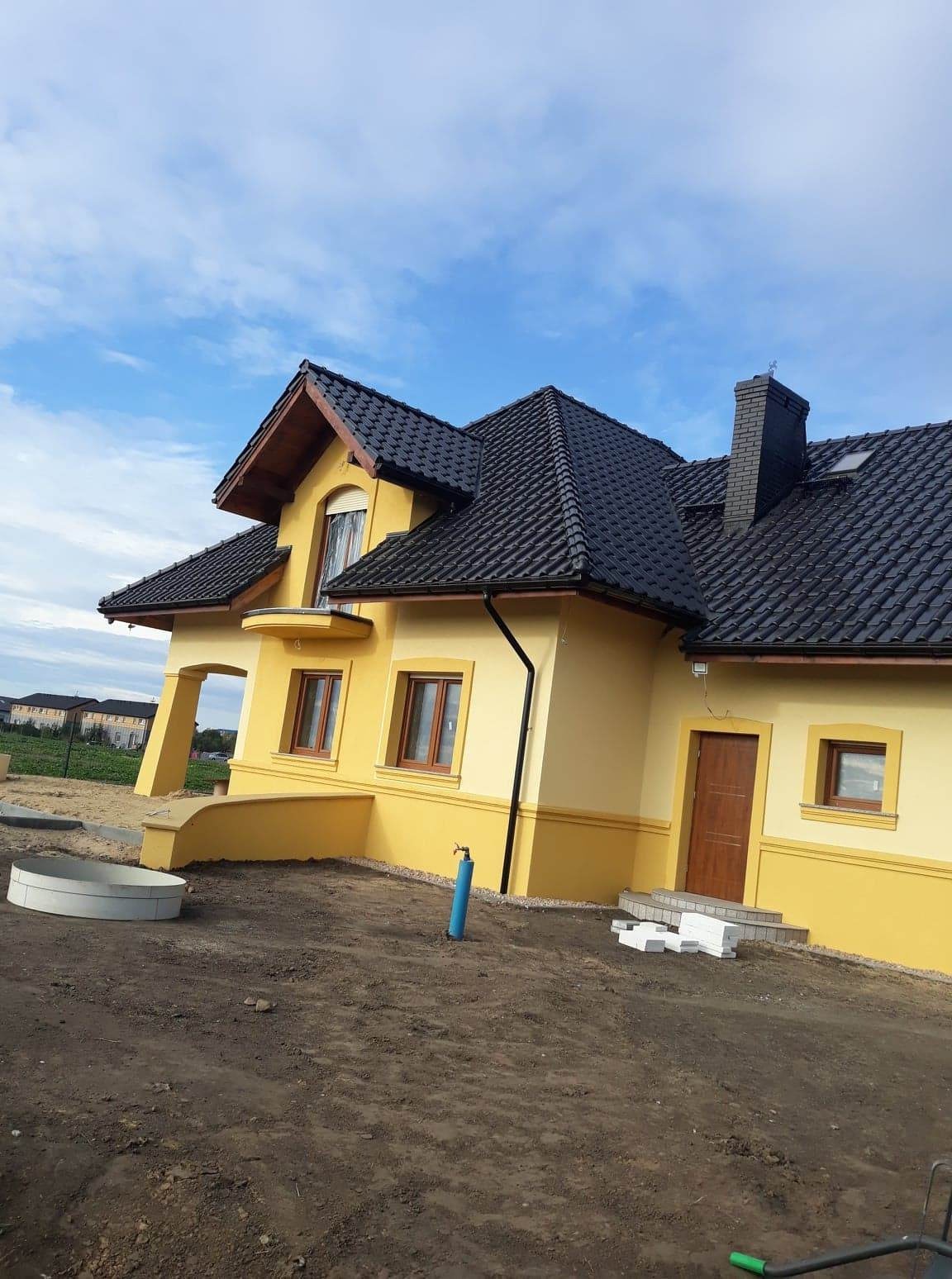 Żółty dom jednorodzinny z ciemną dachówką, widok z boku, teren wokół domu jeszcze niezagospodarowany, widoczne elementy instalacji wodnej i kanalizacyjnej.