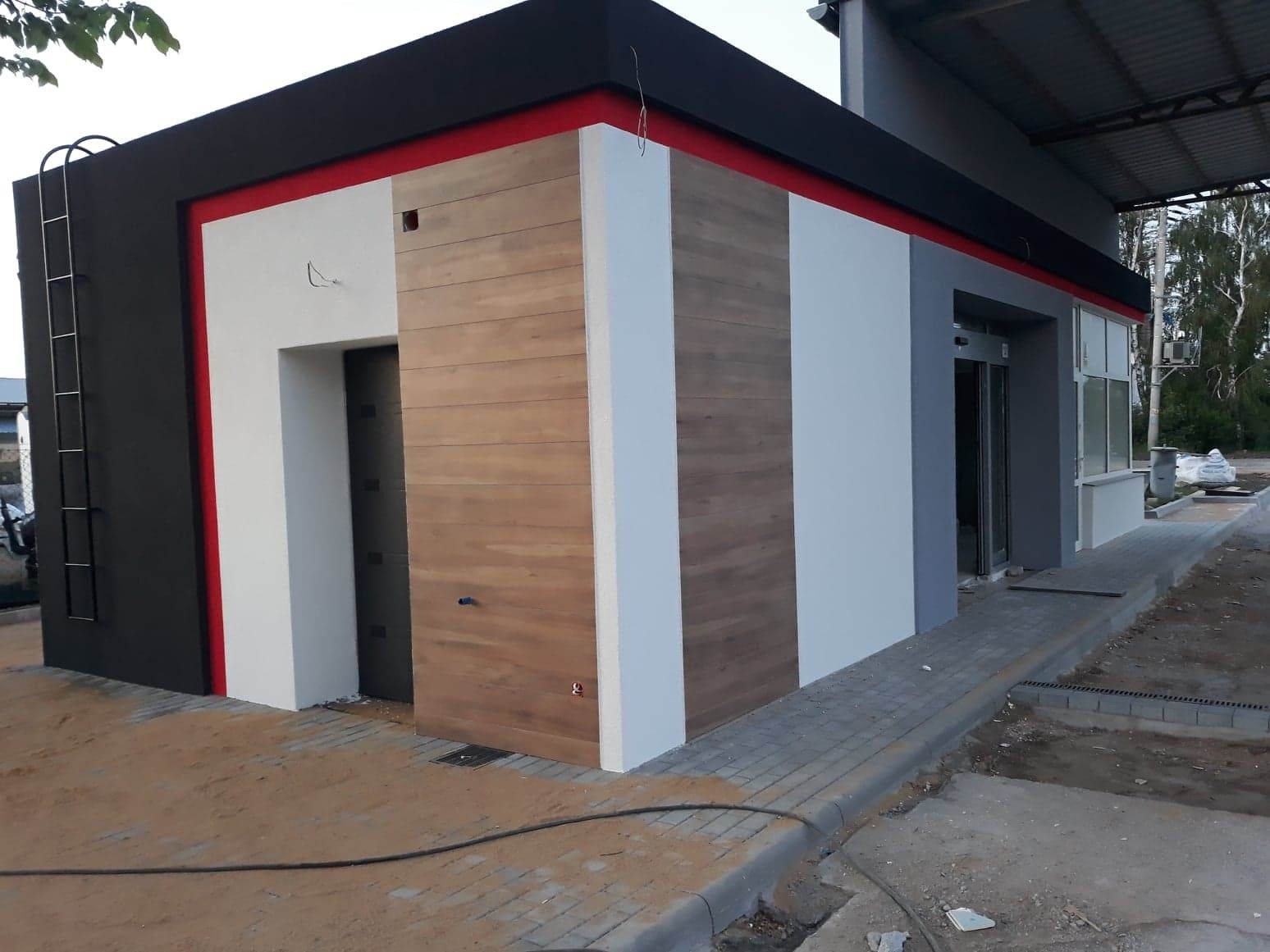 Nowoczesny budynek po ociepleniu i pomalowaniu elewacji na kolory: czarny, czerwony, biały, szary i drewnopodobny, z metalową drabinką na ścianie i automatycznymi drzwiami wejściowymi.