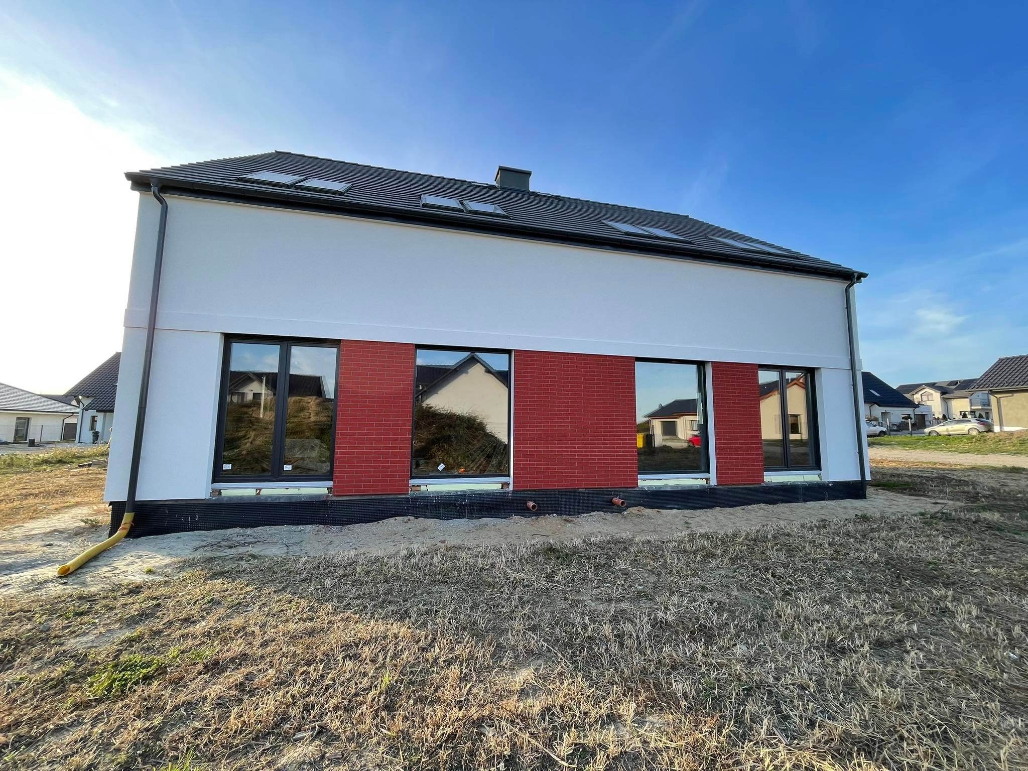 Nowoczesny dom jednorodzinny z białymi ścianami i ceglanym pasem pod oknami, widok z zewnątrz na etapie wykończenia terenu.