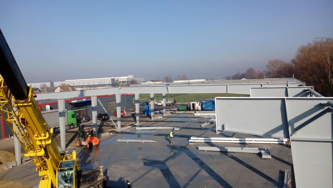 Montaż konstrukcji stalowej hali, widok z góry na plac budowy z dźwigiem, maszynami i pracownikiem w kamizelce odblaskowej.