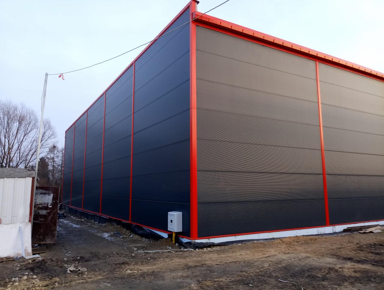 Duży, nowy budynek przemysłowy z czarnymi panelami i czerwonymi elementami konstrukcyjnymi, widoczny z zewnątrz na nieutwardzonym terenie.